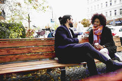 Man and woman with drinks sitting on a bench outside while having a conversation.