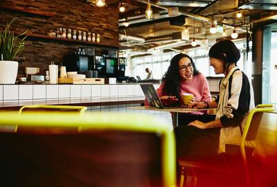 two females working and smiling on their laptops in a cafe