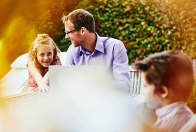man smiling with a child next to him while working on a laptop outside