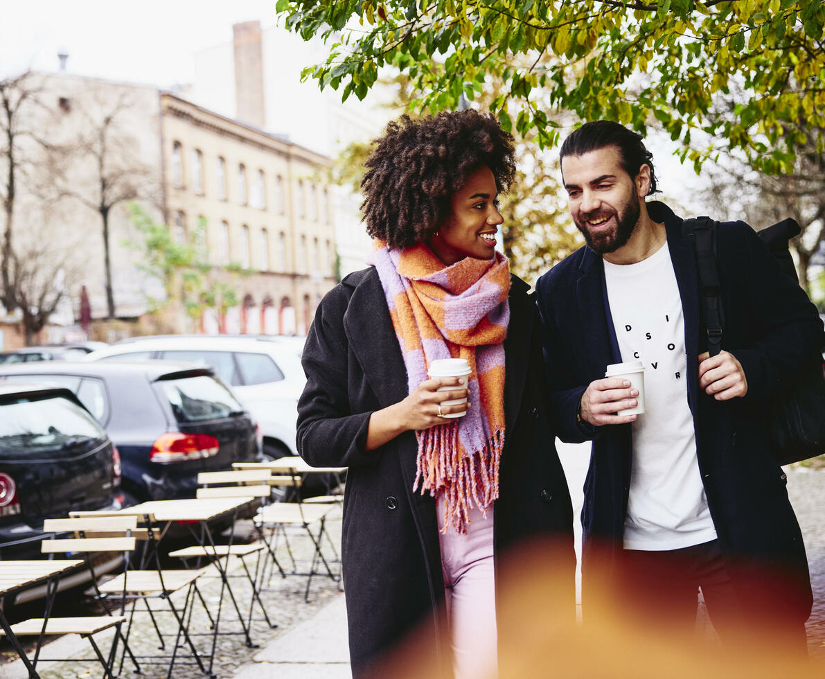 Smiling male and female having a conversation while holding drinks and walking outside.

