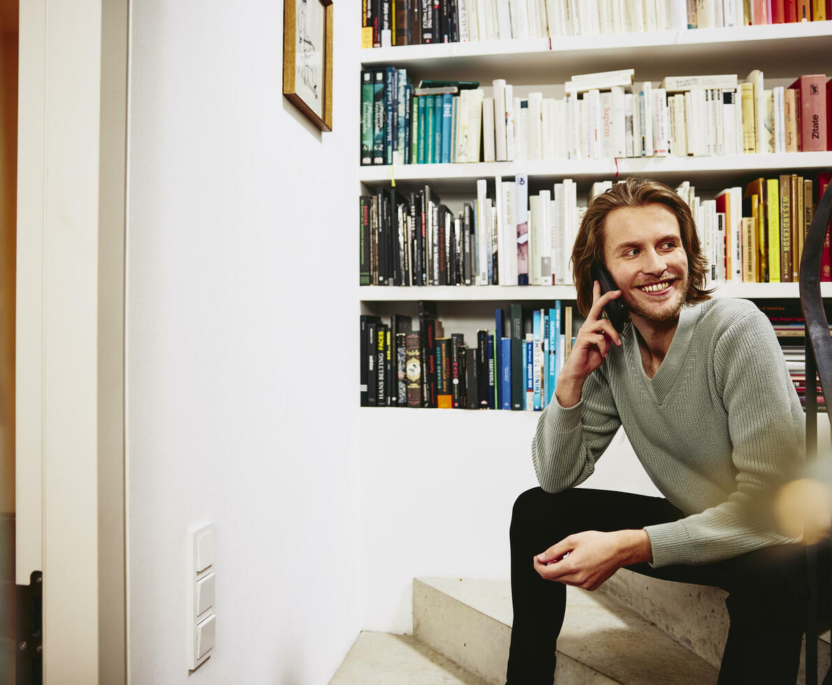 Smiling male sitting on stairs, having a phonecall. bookshelf in background
