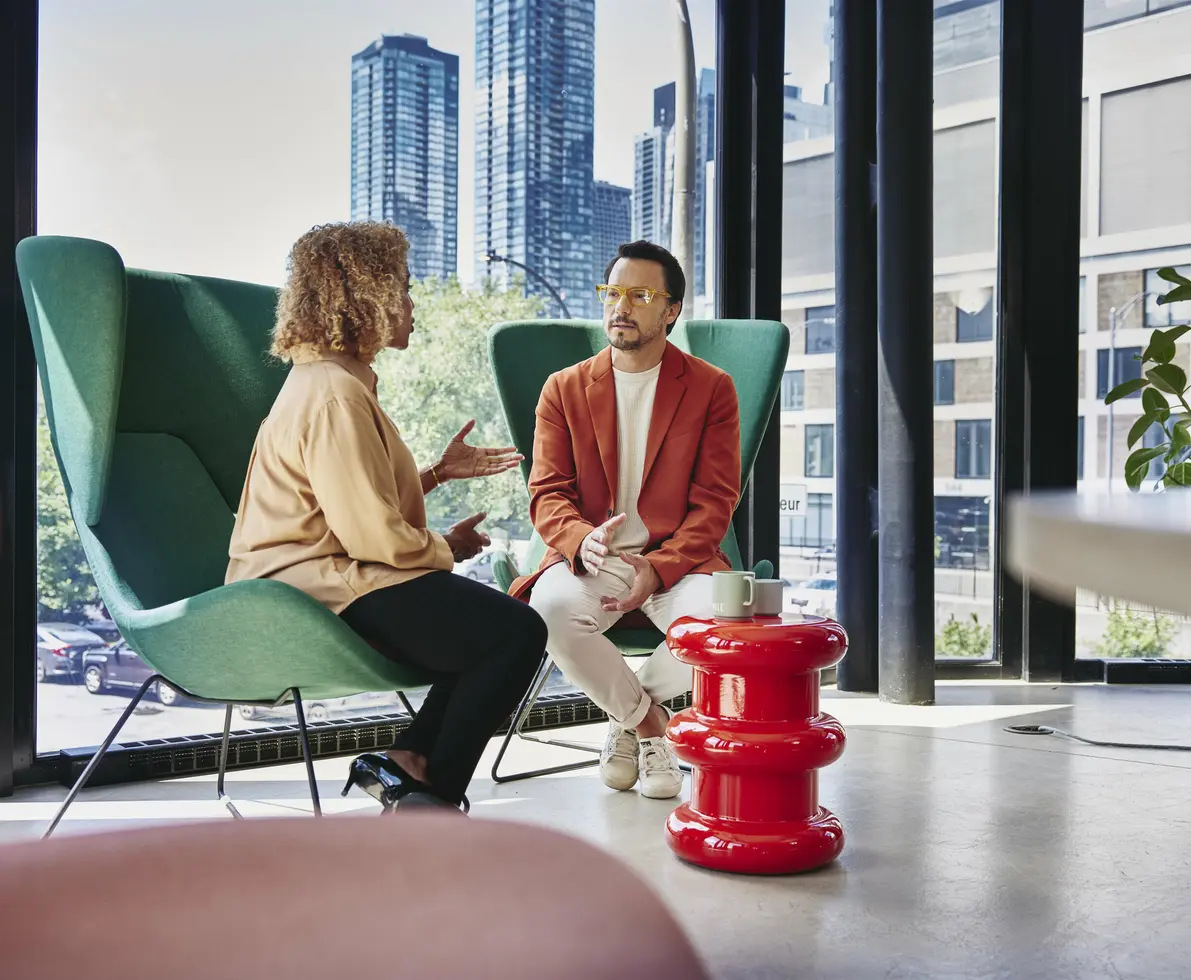 Male with glass and red blazer sitting in a green chair talking to a female sitting in a green chair