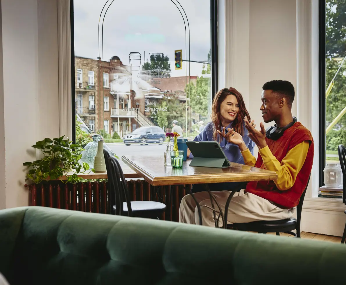 Female smiling towards a male with headphones, sitting next to each other at a table with standing tablet, in a cafe