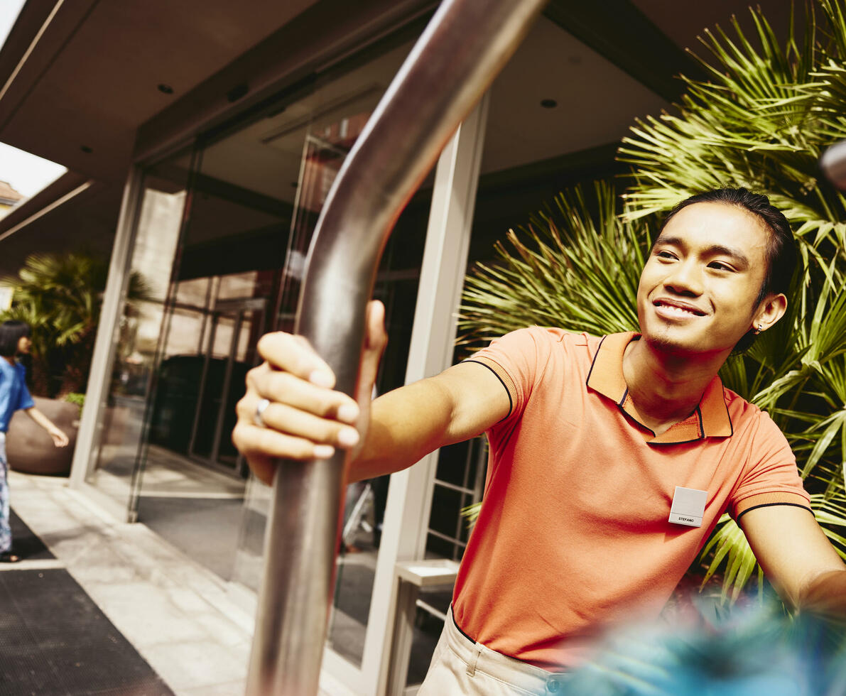 Smiling bellhop pushing a luggage trolley outside a hotel entrance.
