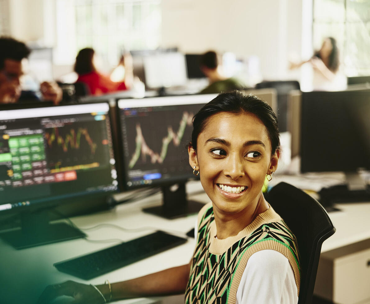 Smiling female looking away from computer screens displaying financial information.
